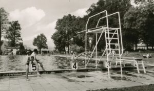 Schwimmbecken des Freibads im Stadtpark in schwarz-weiß mit einem 3-Meter-Brett im Vordergrund und Menschen im Wasser und im Gras