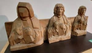 drei geschnitzte Holzfiguren in unterschiedlichen Schnitzstadien, von der groben Silhouette über etwas detailreicher bis hin zur fertigen Figur