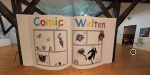 Blick auf die Eingangsinstallation der Ausstellung "Comic-Welten". Ein Menschenhohes, aufgeschlagenes Comicheft mit verschiedenen Comic-Figuren: Snoopy, Spiderman, Tim und Struppi, Pokemon, Son Goku und andere